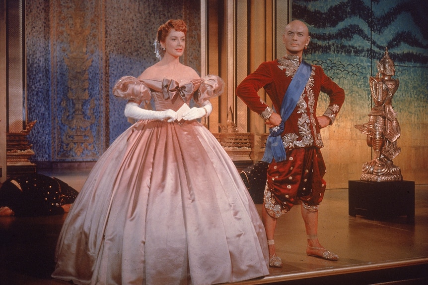 A 1950s still from a film with an English woman in a pink gown and a man dressed as a Thai king with a red outfit and blue sash.