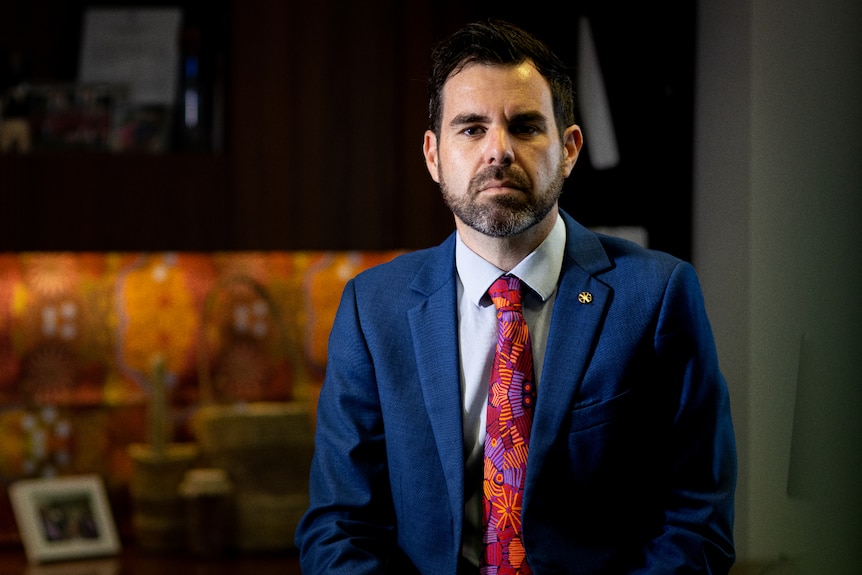 NT government Minister stands in a blue suit and red tie in his office looking concerned.