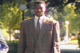 Kendal 'Tiny' Pinder walks with his head downcast wearing a brown suit along an outdoor path.