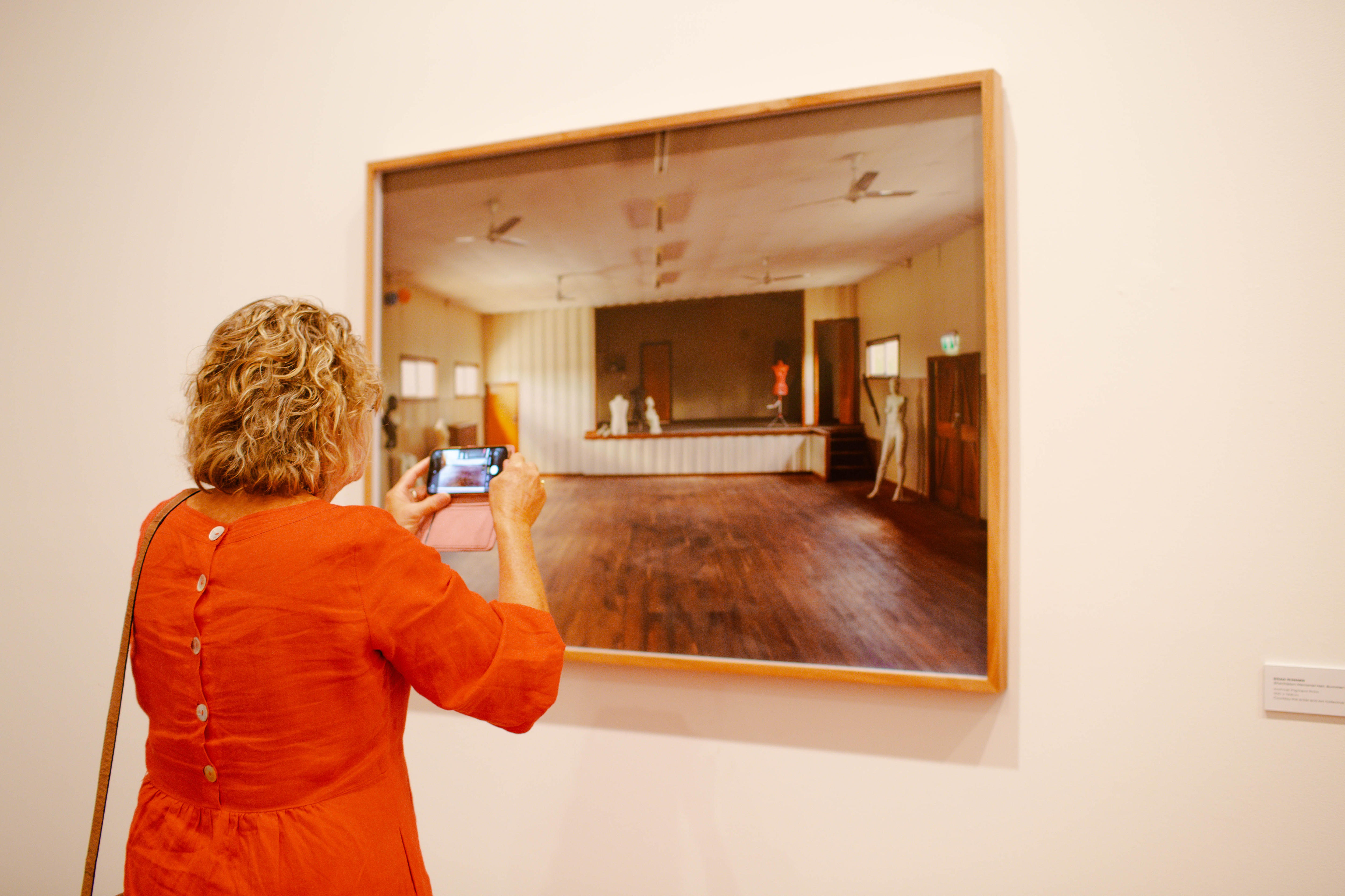 Woman wearing orange dress uses phone to photograph a photograph hanging on gallery wall 