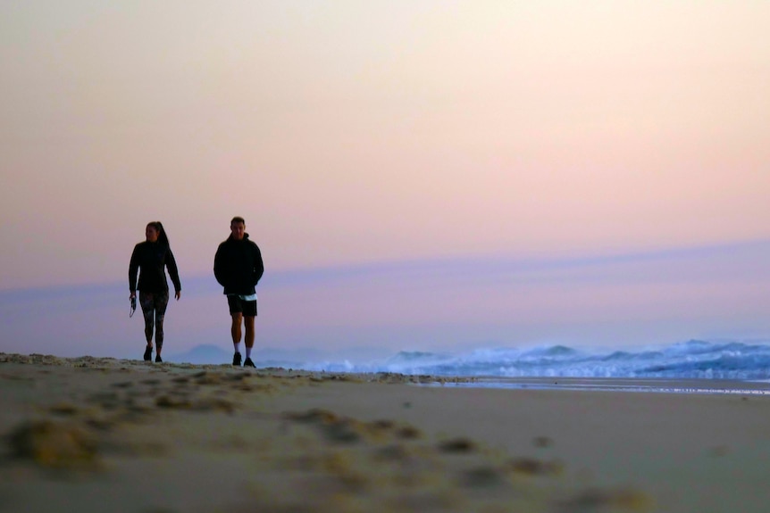 A man and a woman walk on beach at sunrise. They are almost silhouetted by the sky behind them.
