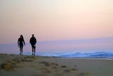 A man and a woman walk on beach at sunrise. They are almost silhouetted by the sky behind them.