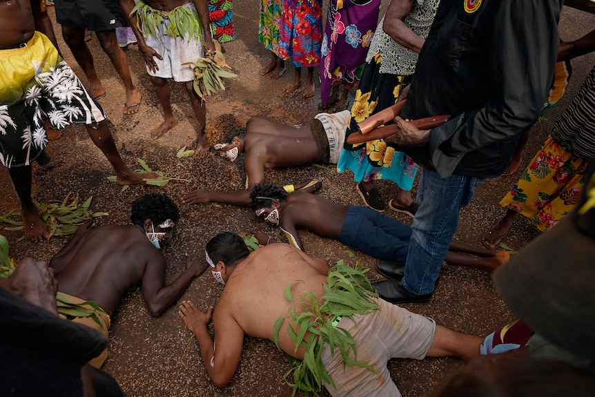 Four men with painted faces lay on the floor. They are surrounded by people.