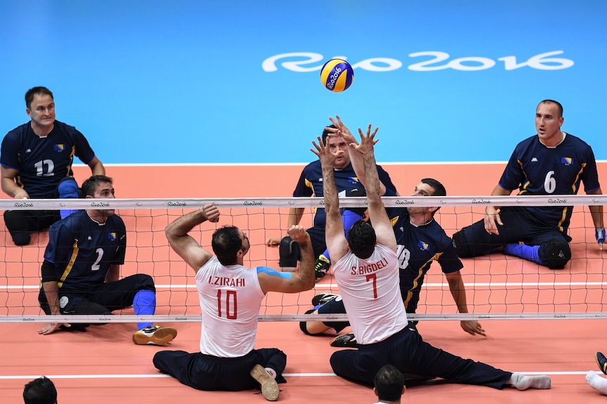 A group of sitting volleyballers raise their arms at the net to block a ball.