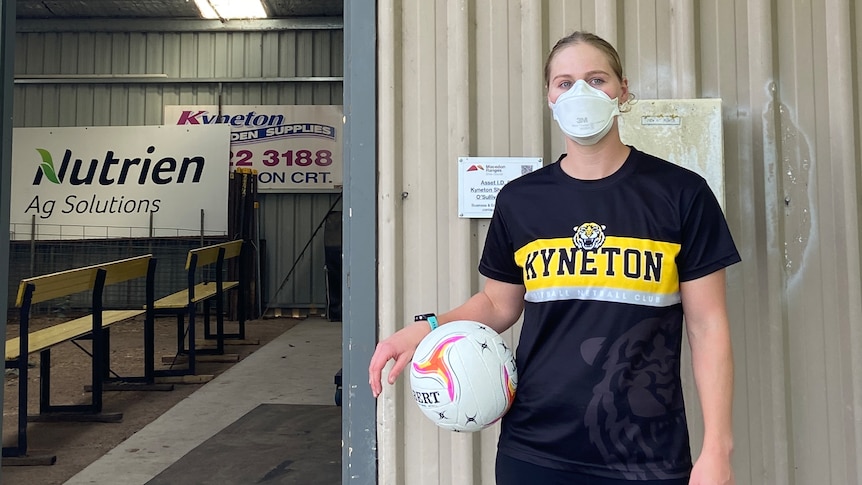 a photo of a woman standing in front of the entrance to a shed with a netball in her hand and a face mask on