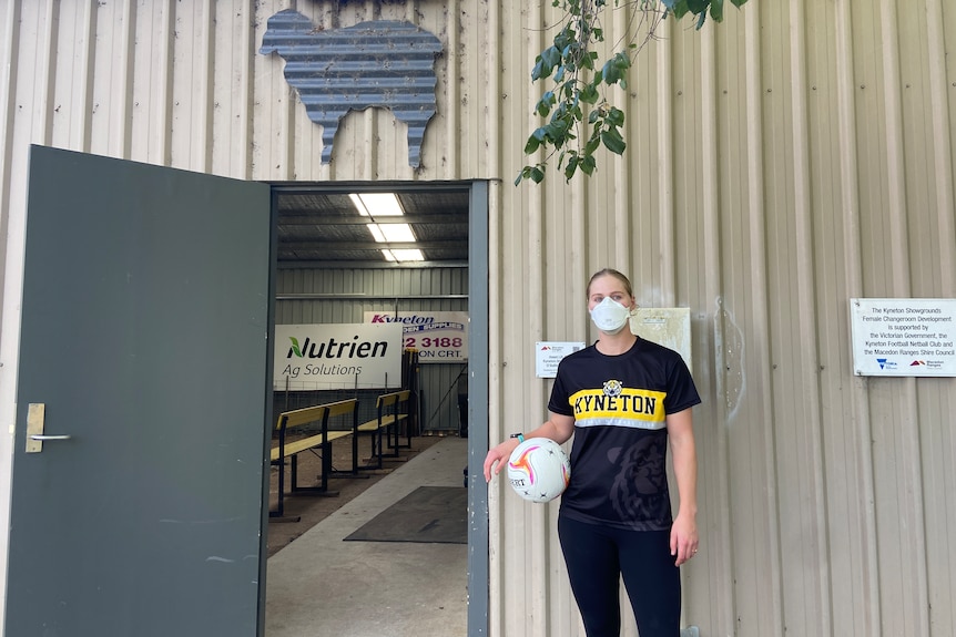 a photo of a woman standing in front of the entrance to a shed with a netball in her hand and a face mask on