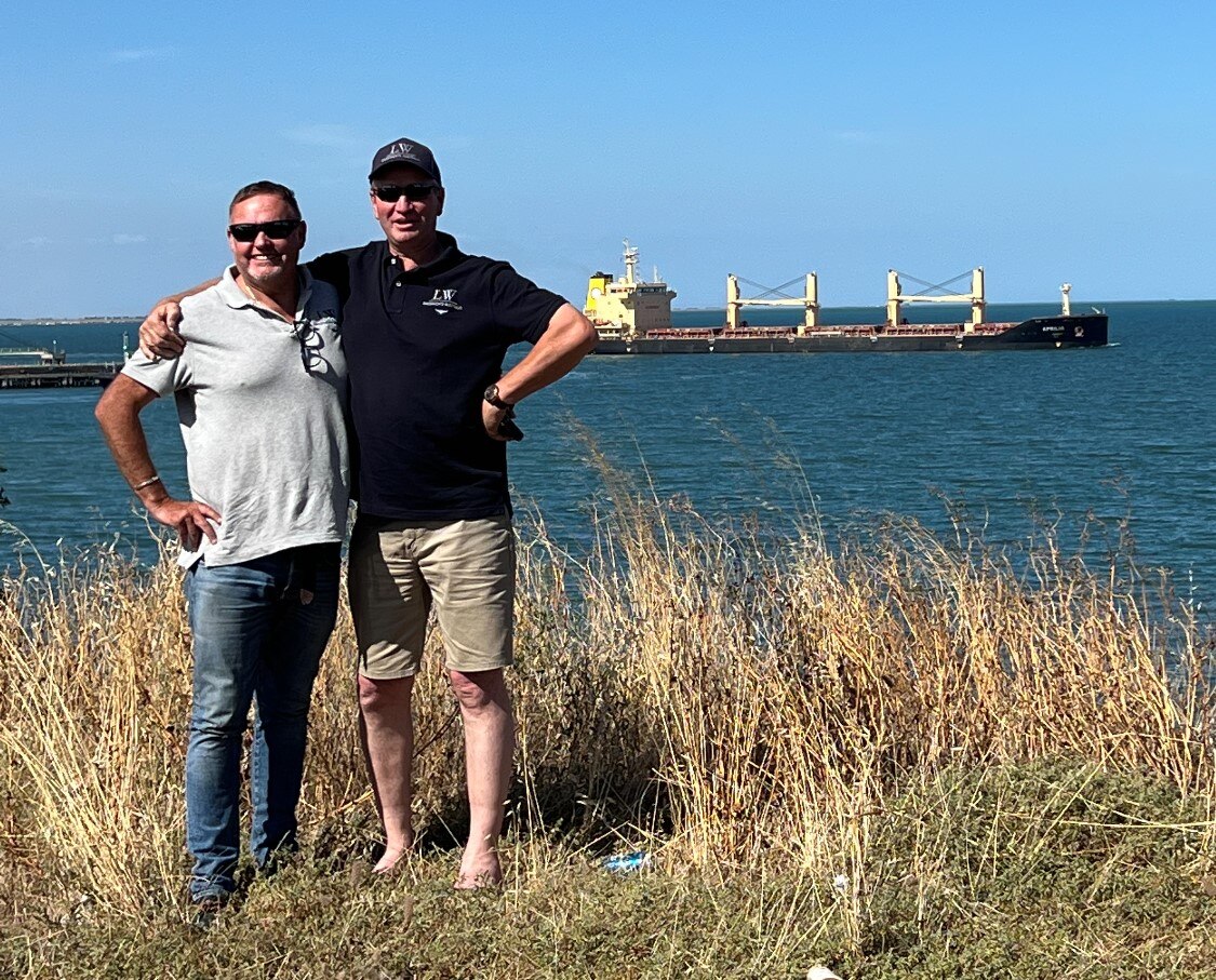 Two men stand with a ship behind them 