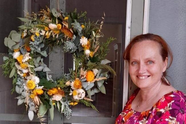 A lady standing next to a door that has a floral wreath hanging on it 