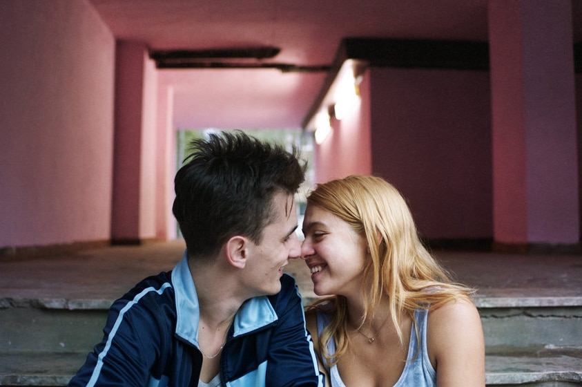 A young man with dark short hair and young woman with long blonde hair sit on exterior steps near pink corridor and touch noses.