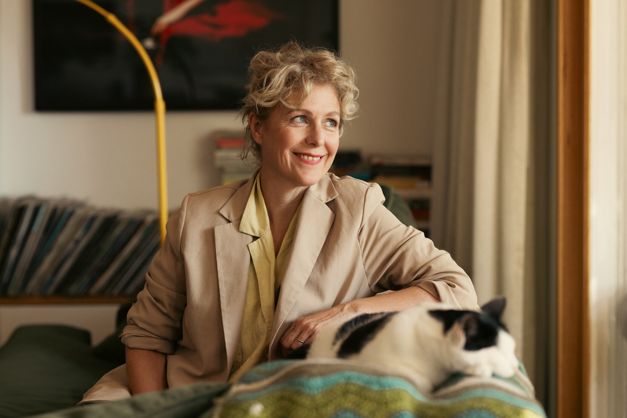 A person with fair curly hair sits on a couch looking out a window smiling, with a cat near her arm.