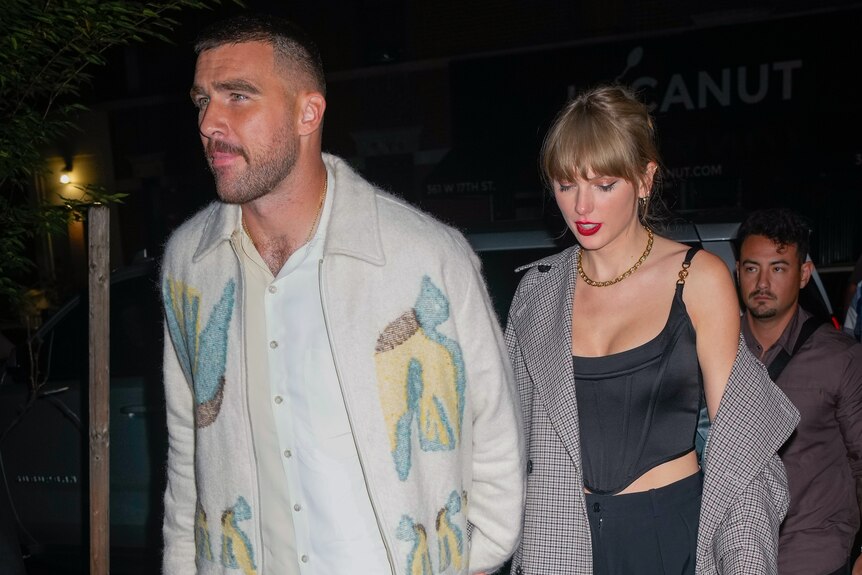 Taylor Swift wearing loose shirt over black corset holds hands with Travis Kelce as they walk outside at nighttime