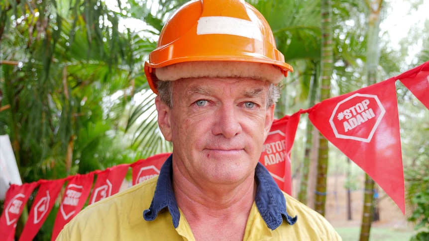 A man wearing a hard hat and high vis shirt stand in front of red flags that read "stop Adani".