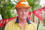 A man wearing a hard hat and high vis shirt stand in front of red flags that read "stop Adani".