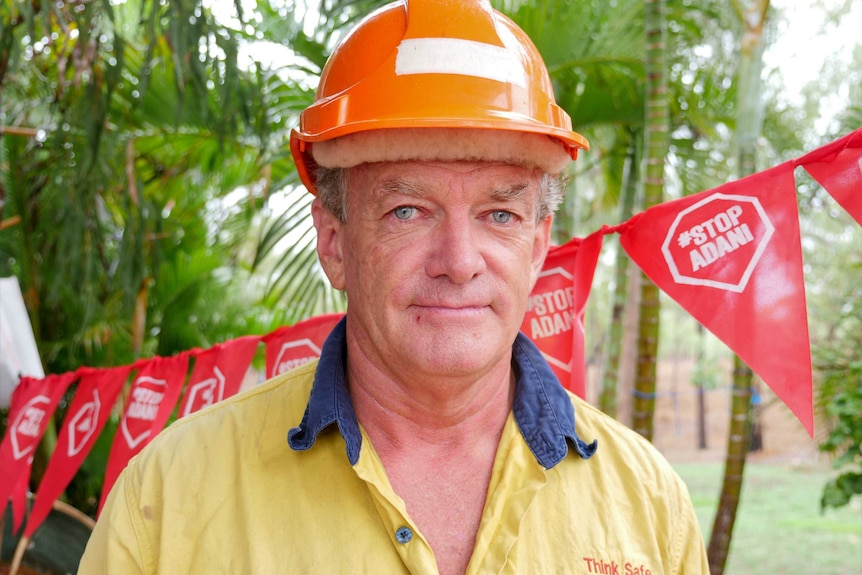 A man wearing a hard hat and high vis shirt stand in front of red flags that read "stop Adani"