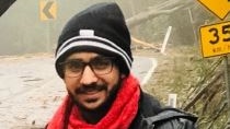 A young man in a black beanie, red scarf and grey pants smiles on a rainy road.