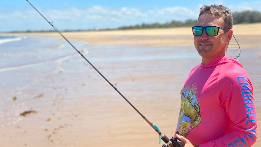 A man in a pink rashie and mirror sunglasses stands on a beach holding a fishing rod.