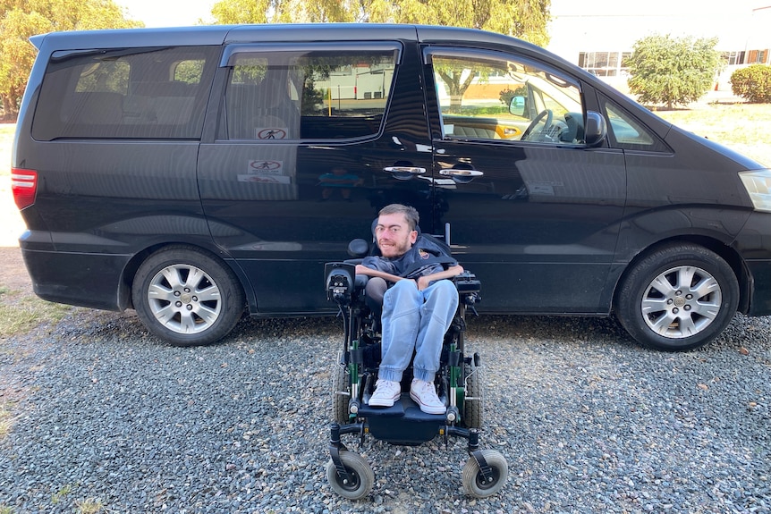 A smiling young man in a motorised wheelchair sits in front of a black van, wears jeans, white sports shoes.