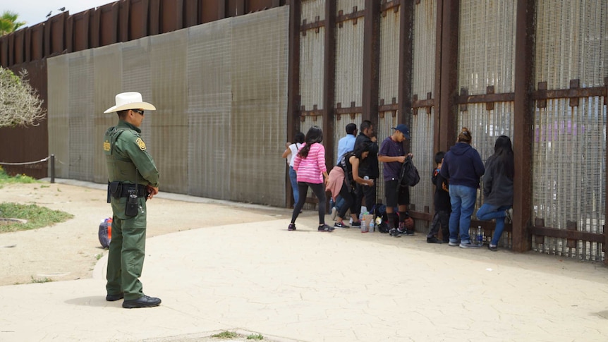 An agent watches as people on the US side of the border speak with friends and family in Mexico through a mesh fence.