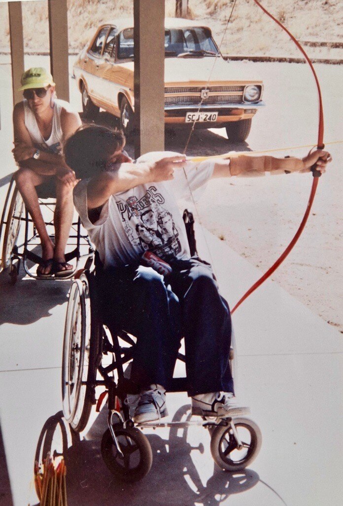 young man with brown hair, white tshirt in wheelchair plays archery ready to fire