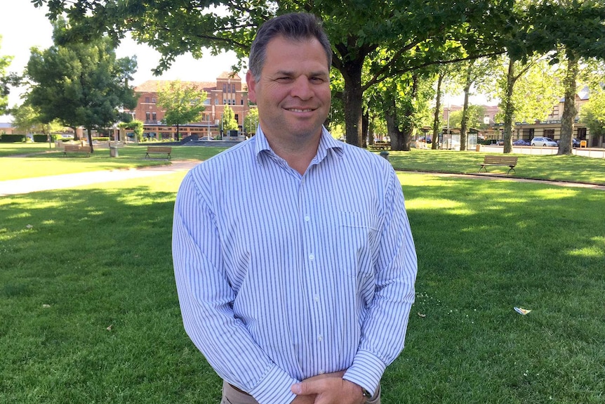 Man stands in leafy park with hands folded together. He is wearing an unbuttoned striped blue and white shirt and chinos.