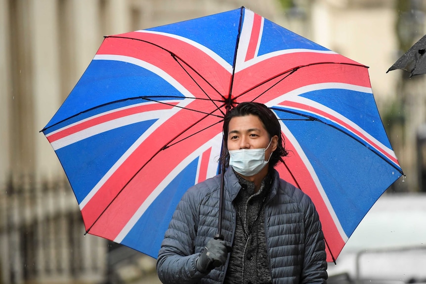 A man wearing a face mask and holding an umbrella with the Union Jack on it waits outside 10 Downing Street.