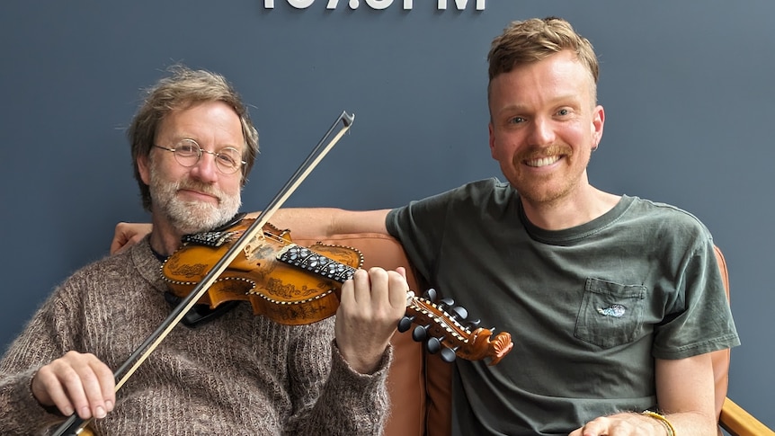 Musician Martin Scuffin sits on a couch holding his fiddle next to ABC broadcaster Matt Tribe. 