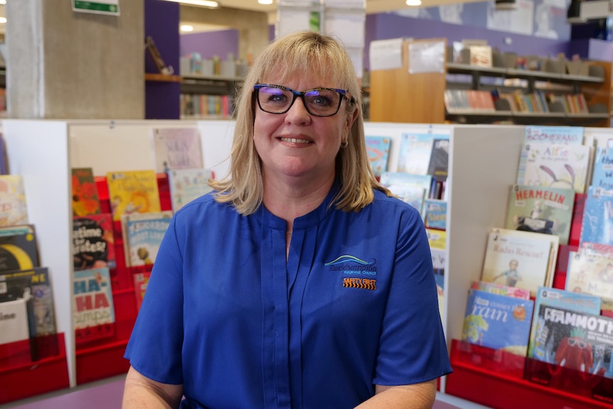 A woman smiles at the camera, in the background are shelves of picture books. 