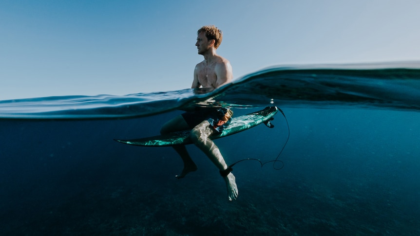 An over/under photo of Brett Connellan, shirtless and sitting on a surfboard in the ocean.