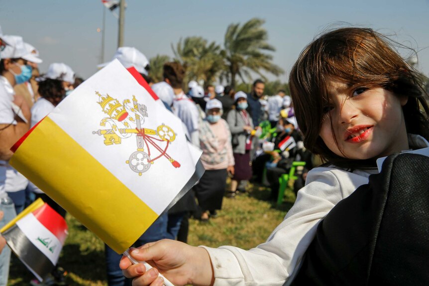 A young girl smiles at camera and carries a white, yellow and red flag in a crowd on a sunny day.
