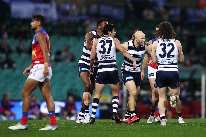 A bald AFL footballer smiles as his happy teammates crowd around him after a goal.