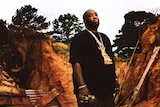 Killer Mike standing outside wearing a black shirt and gold chain, he has a serious expression on his face