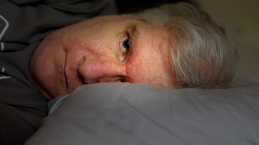 A close up of a man lying in bed with is head on a pillow, his face illuminated by light while his body is in shadow.