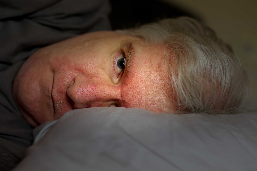 A close up of a man lying in bed with is head on a pillow, his face illuminated by light while his body is in shadow.