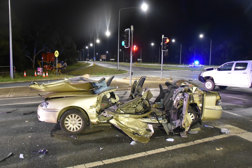 A sedan with its roof cut off and significant damage at an intersection, with a police car in the background.