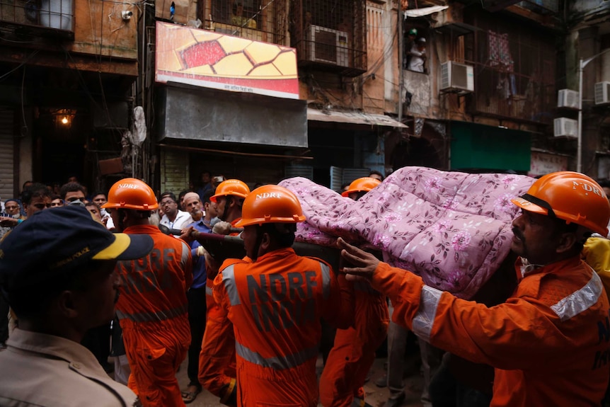 A group of rescue men clad in orange jumpsuits carry a deceased body wrapped in a pink blanket decorated with flowers.