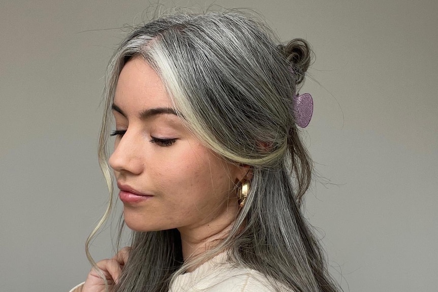A young woman with shiny grey hair in a half-updo looks over her shoulder