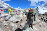 A young boy stands in front of a line of prayer flags with mountains in the distance. 