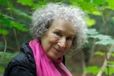Author Margaret Atwood standing in a forest smiling and looking at the camera.