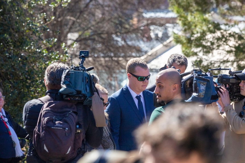 A man in sunglasses stares blankly at a media scrum with TV cameras.