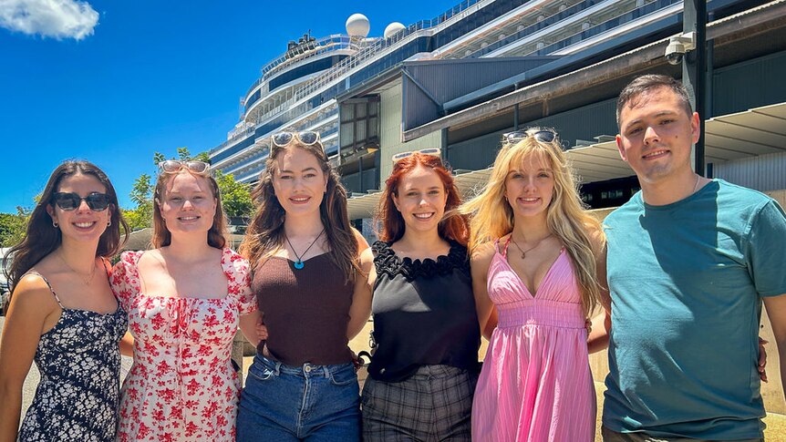 A group of young people stand arm-in-arm in front of a cruise ship terminal.