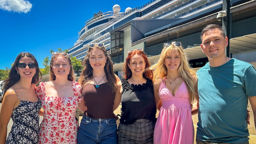 A group of young people stand arm-in-arm in front of a cruise ship terminal.