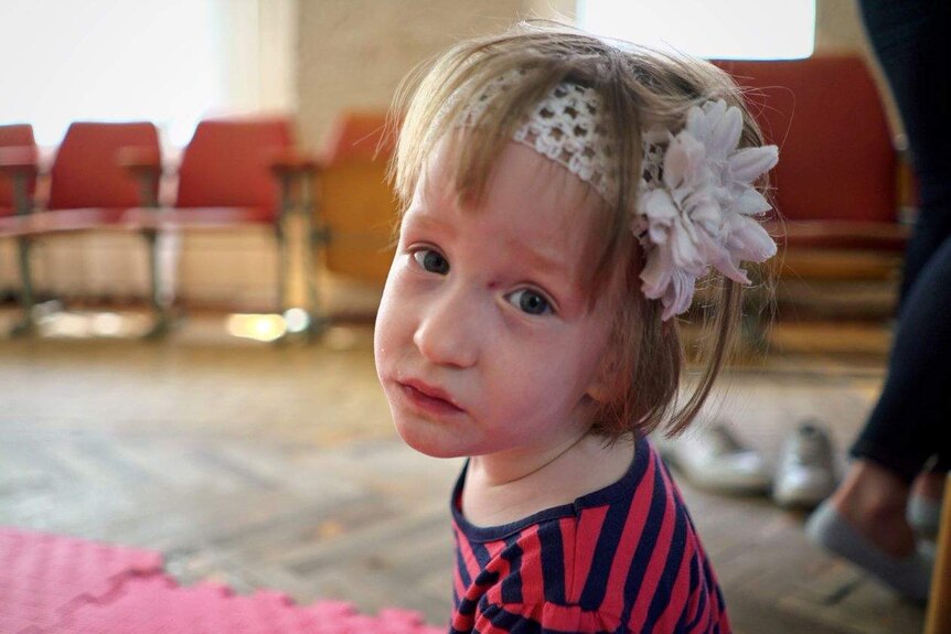 A three-year-old girl wearing a white headband sits on rubber mats on the floor, looking at the camera with a sad expression.