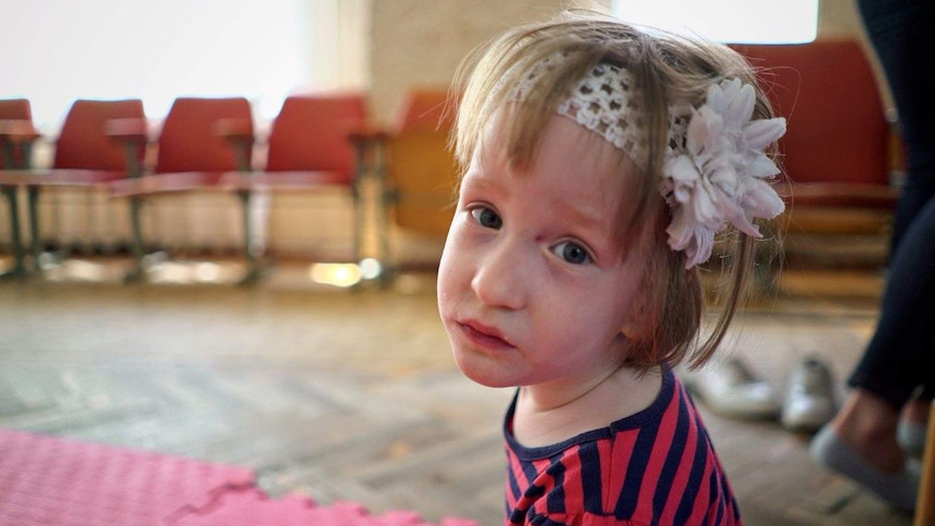 A three-year-old girl wearing a white headband sits on rubber mats on the floor, looking at the camera with a sad expression.