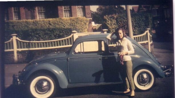 A woman stands in front of a blue VW Beetle.