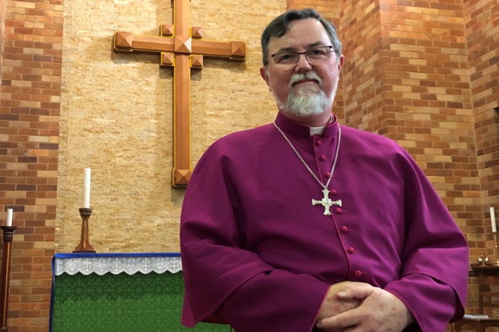 Bishop Donald Kirk in his purple vestments stands in front of the altar and a wooden cross hanging on the wall.