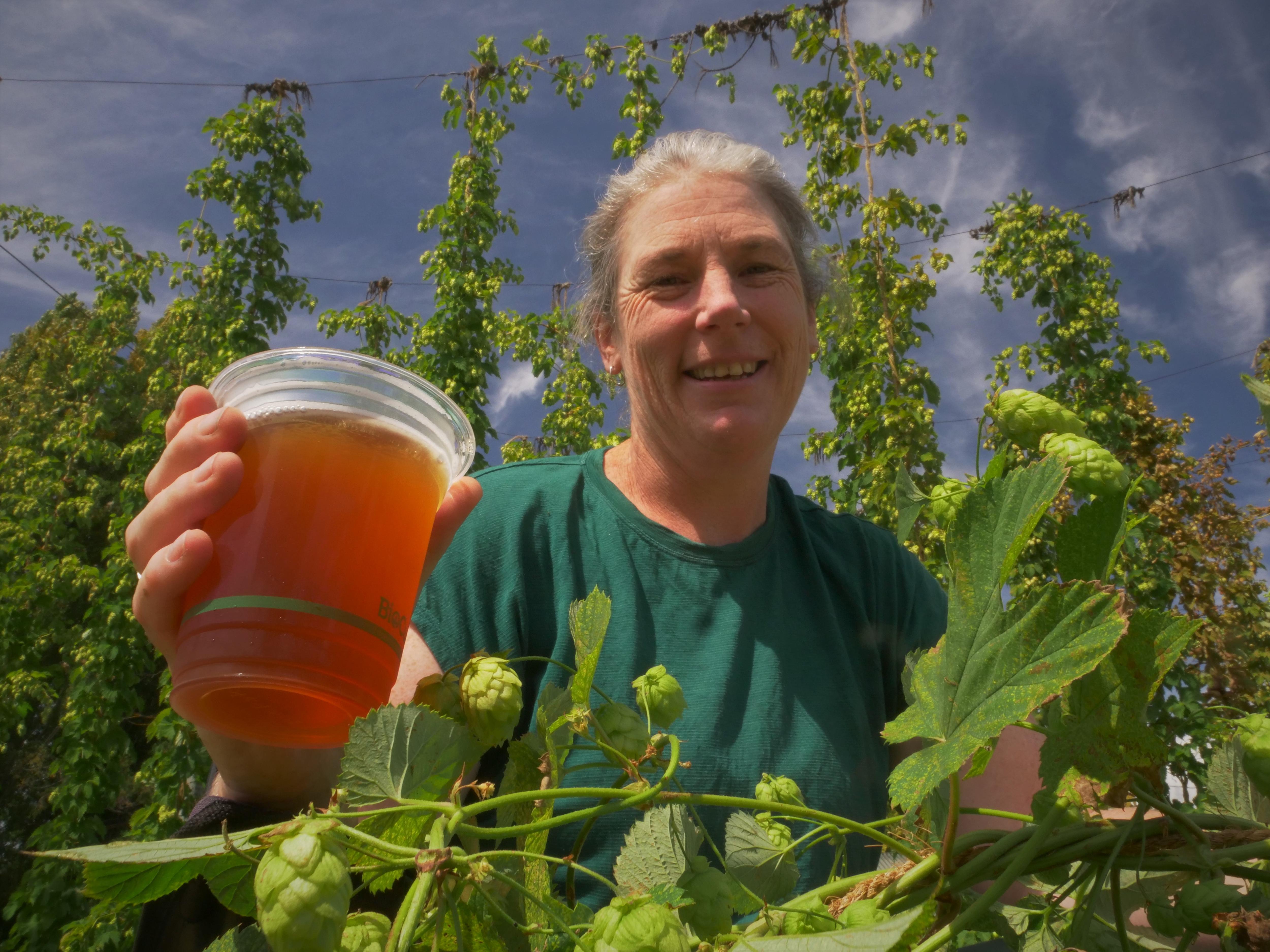 A woman smiling as she holds a beer up in the sun, hop vines in the foreground.