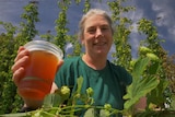 A woman smiling as she holds a beer up in the sun, hop vines in the foreground.