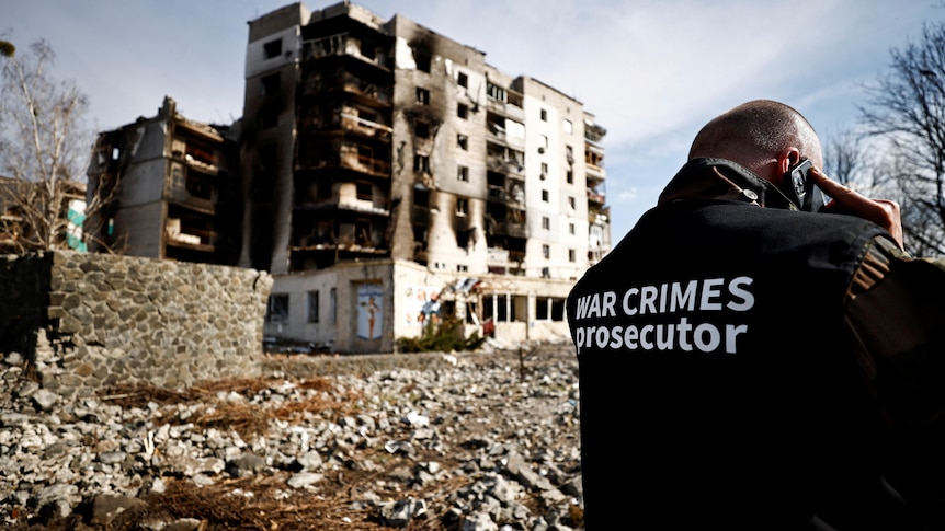 A man in a jacket with text saying 'war crimes prosecutor' stands in front of a burned building