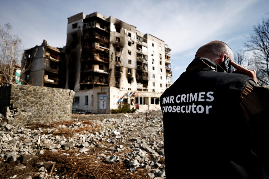 A man in a jacket with text saying 'war crimes prosecutor' stands in front of a burned building