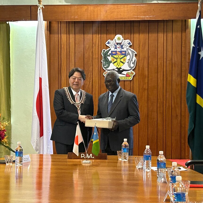 Japanese foreign minister and Solomon Islands PM hold white box and smile at end of table.